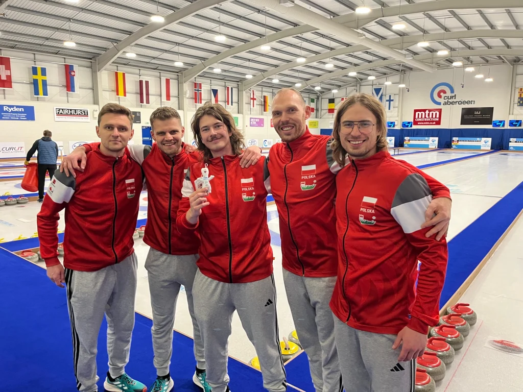 Męska reprezentacja Polski w curlingu Pięciu mężczyzn ubranych w czerwone sportowe kurtki polskiej reprezentacji stoi razem na hali do curlingu, uśmiechają się i pozują do zdjęcia, jeden z nich trzyma w ręku maskotkę.
