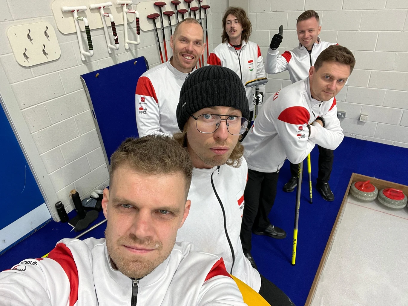 Sześciu mężczyzn ubranych w biało-czerwone kurtki drużynowe pozuje na arenie curlingowej, na ścianie za nimi wiszą miotły do curlingu, a na lodzie widoczne są kamienie curlingowe.