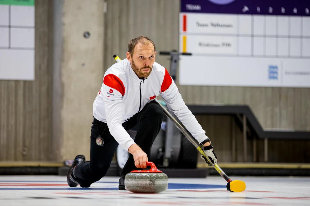 Bartosz Łobaza Mężczyzna w sportowym stroju pochylony nad lodową taflą, pcha przed siebie curlingowy kamień, trzymając w drugiej ręce szczotkę do curlingu, w tle widoczne plansze wyników.