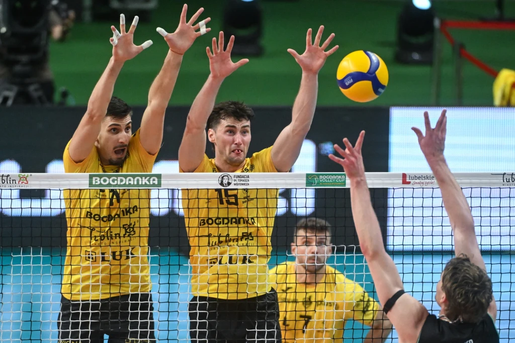 Dwóch zawodników w żółtych koszulkach unosi ręce do bloku nad siatką, naprzeciwko zawodnik w czarnej koszulce szykuje się do ataku, piłka jest nad siatką, w tle widoczni kibice i hala sportowa.
