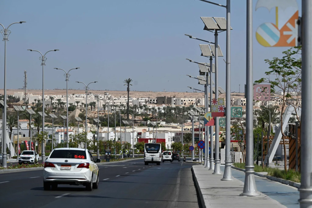 Tragiczny wypadek katarskich dyplomatów miał miejsce przed szczytem odbywającym się w egipskim Szarm el-Szejk (na zdjęciu) Ruchliwa ulica miejska z kilkoma samochodami oraz autobusem poruszającymi się w obu kierunkach, po bokach chodniki z latarniami oraz banery dekoracyjne, w tle panorama zabudowań miejskich i pojedyncze palmy na wzgórzu.
