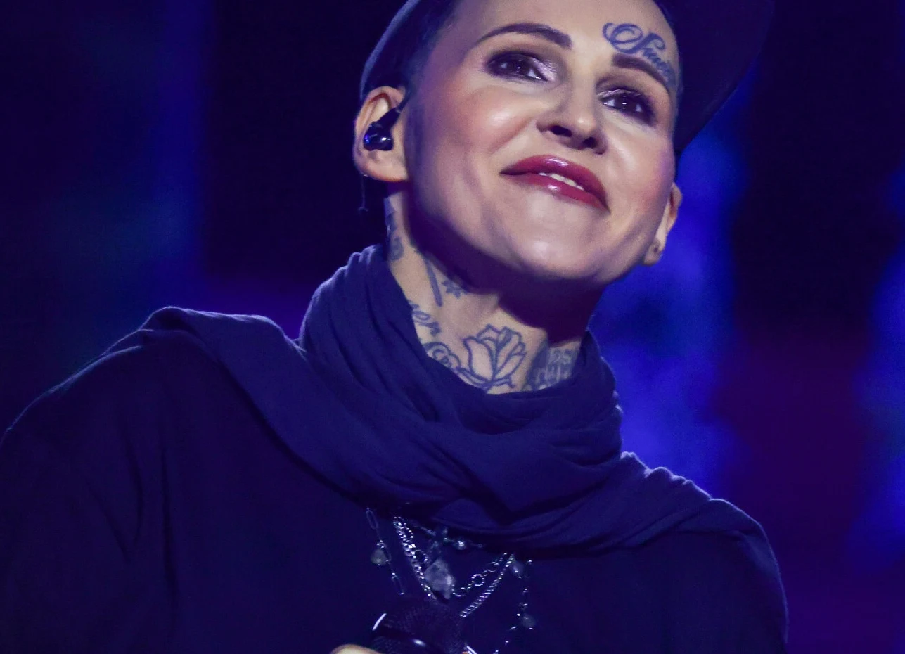 Agnieszka Chylińska zakończyła trasę po USA Osoba z tatuażami na twarzy i szyi, uśmiechnięta, ubrana w ciemne ubrania, stoi na tle rozmytego, kolorowego światła.