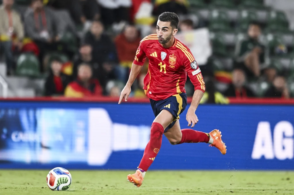 Piłkarz w czerwonym stroju Hiszpanii biegnie z piłką po murawie podczas meczu, skupiony na grze, wokół rozmyte sylwetki kibiców na trybunach.