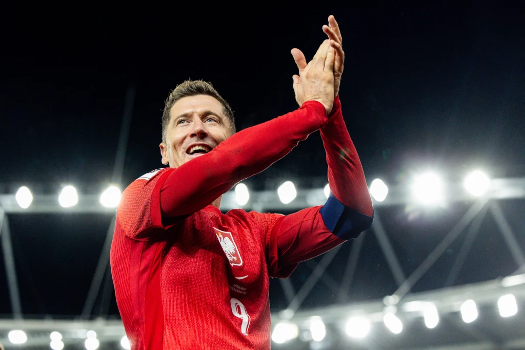 Robert Lewandowski Piłkarz w czerwonym stroju z numerem 9, z opaską kapitana na ramieniu, stoi na stadionie i uśmiecha się, klaszcząc w dłonie, w tle jasne światła stadionowe.