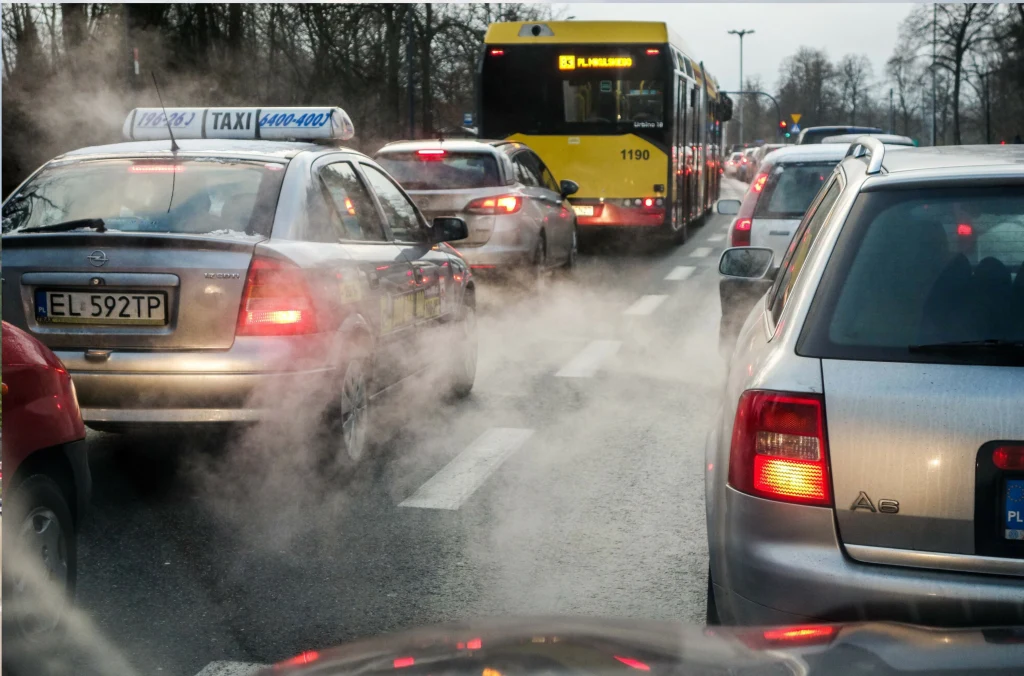 Gęsty korek uliczny, sznur samochodów w dwóch pasach, widoczny autobus miejski oraz taksówka z widocznym znakiem TAXI, unoszące się spaliny z samochodów, ponura, zimowa aura.
