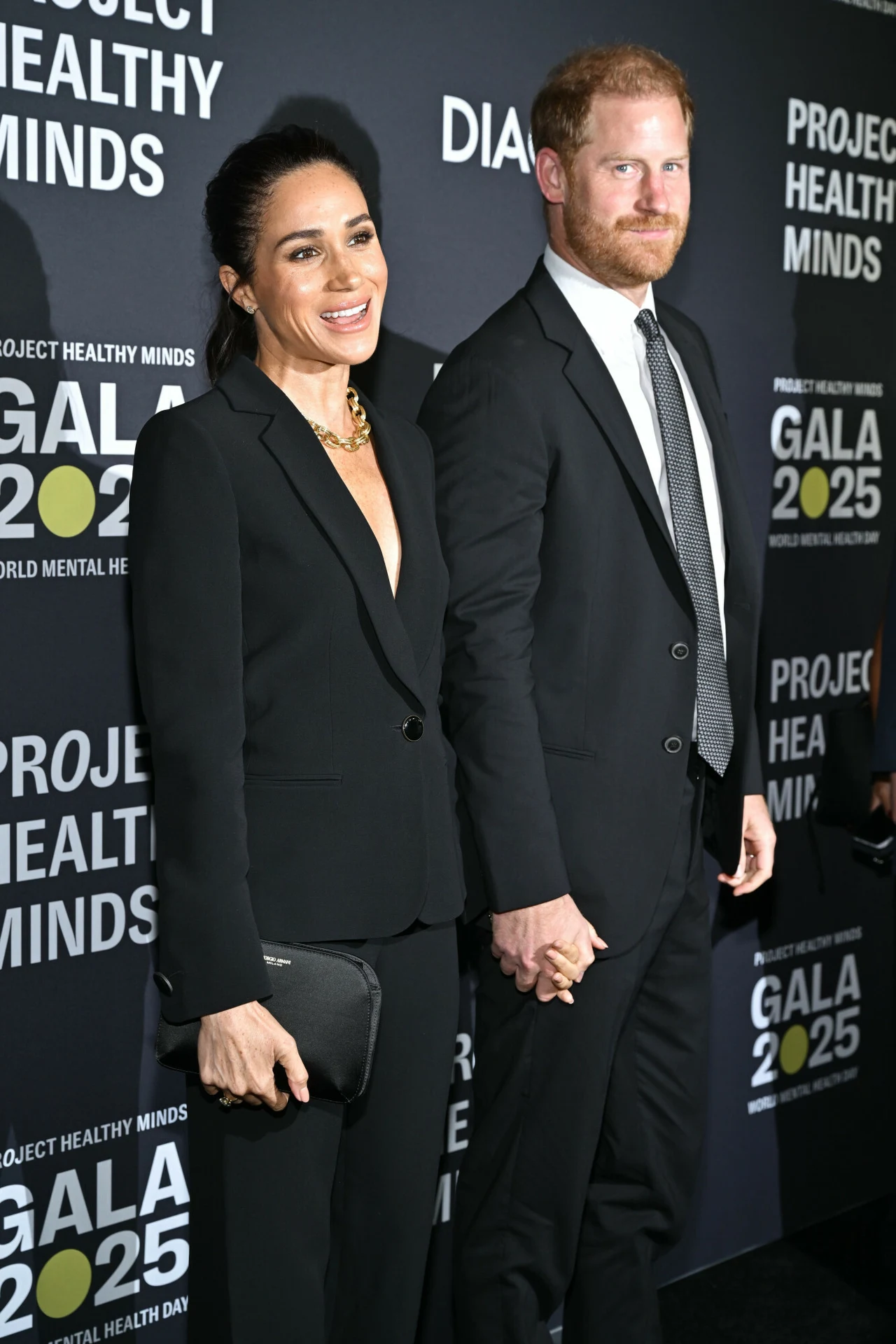 Monochromatyczne i minimalistyczne stylizacje to znak rozpoznawczy Meghan Markle Elegancka para ubrana w czarne garnitury pozuje razem, trzymając się za ręce na tle ścianki z napisem 'Project Healthy Minds' oraz 'Gala 2025'. Kobieta trzyma kopertówkę, oboje uśmiechnięci, sprawiają wrażenie zadowolonych.