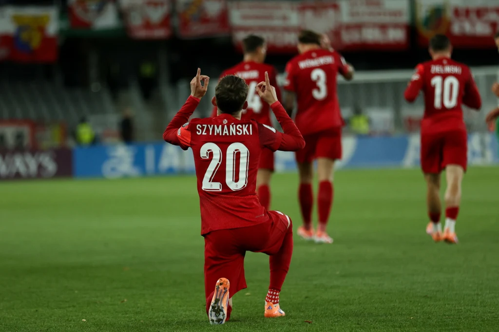 Sebastian Szymański Piłkarz w czerwonym stroju z numerem 20 i nazwiskiem Szymański klęczy na boisku z uniesionymi palcami w geście triumfu, podczas gdy kilku innych zawodników w podobnych strojach znajduje się w tle, idąc razem po murawie stadionu.
