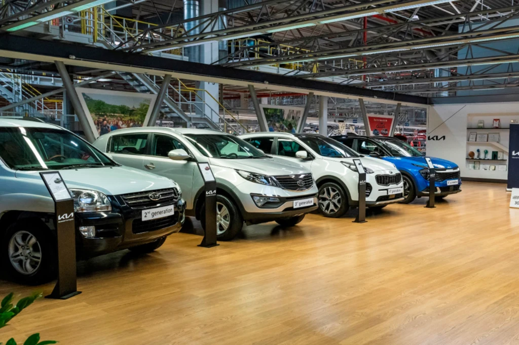 Produkcja Sportage drugiej generacji w zakładzie w Żylinie rozpoczęła się w 2007 r. Cztery nowoczesne samochody osobowe ustawione w rzędzie na wystawie w salonie samochodowym, wyraźnie widoczne modele i szyldy przy każdym pojeździe, w tle elementy hali wystawienniczej.