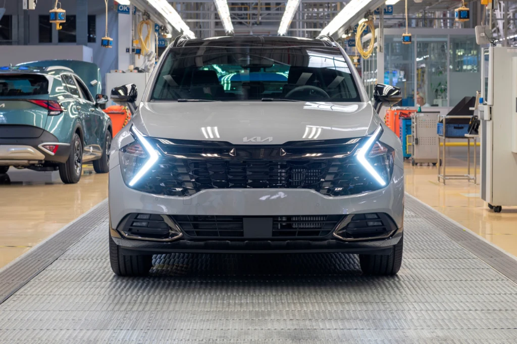 To ostatni proces, podczas którego odpalany jest samochód. Po przejechaniu trafia na szczegółową kontrolę jakości. Nowoczesny srebrny samochód marki Kia stoi przodem na linii produkcyjnej w hali fabrycznej, z włączonymi światłami do jazdy dziennej w kształcie litery L, w tle widoczne inne samochody oraz elementy wyposażenia zakładu.