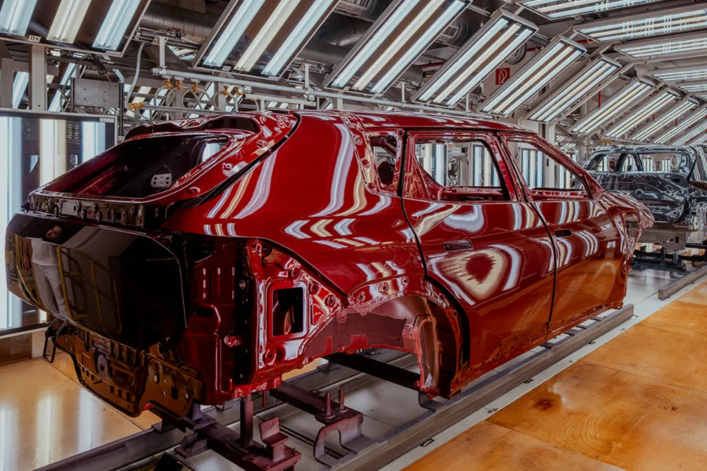 W Żylinie produkowany jest Sportage, Ceed oraz EV4. Czerwona karoseria samochodu na linii montażowej w nowoczesnej fabryce, otoczona silnym oświetleniem przemysłowym, bez zamontowanych okien, kół oraz innych elementów wykończeniowych.