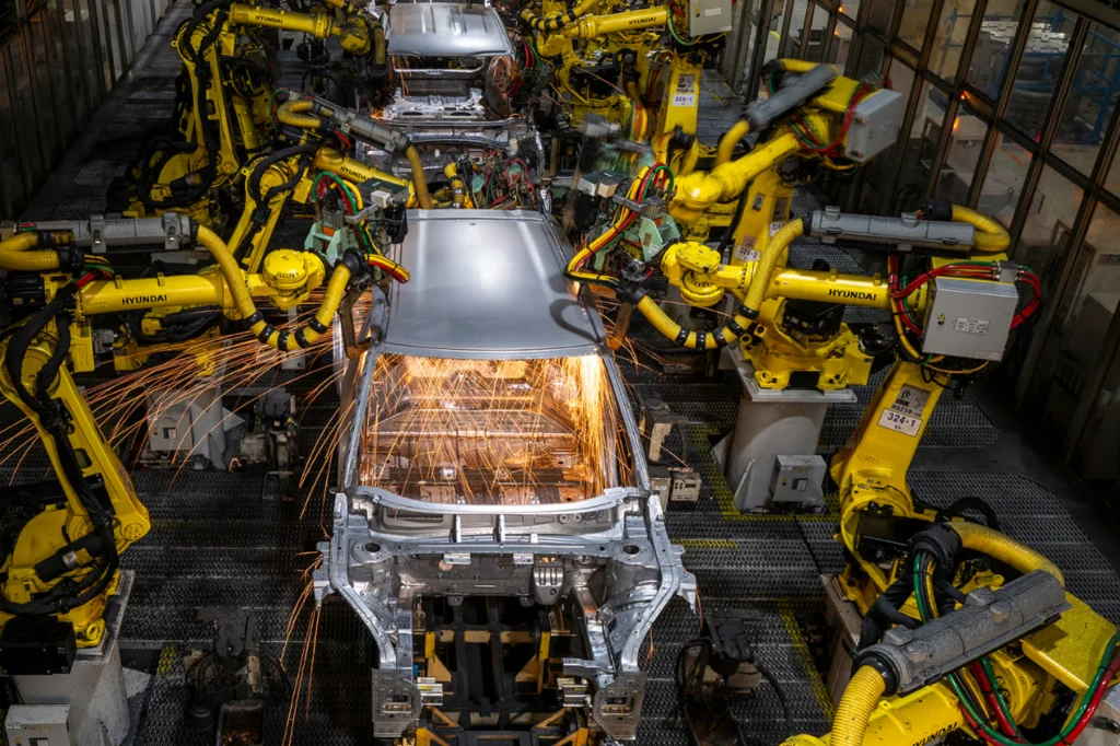 Poszczególne cykle spawania trwają mniej niż minutę. Roboty przemysłowe spawające karoserię samochodu, liczne iskry, stalowa konstrukcja karoserii otoczona żółtymi ramionami maszyn, linia montażowa w fabryce.