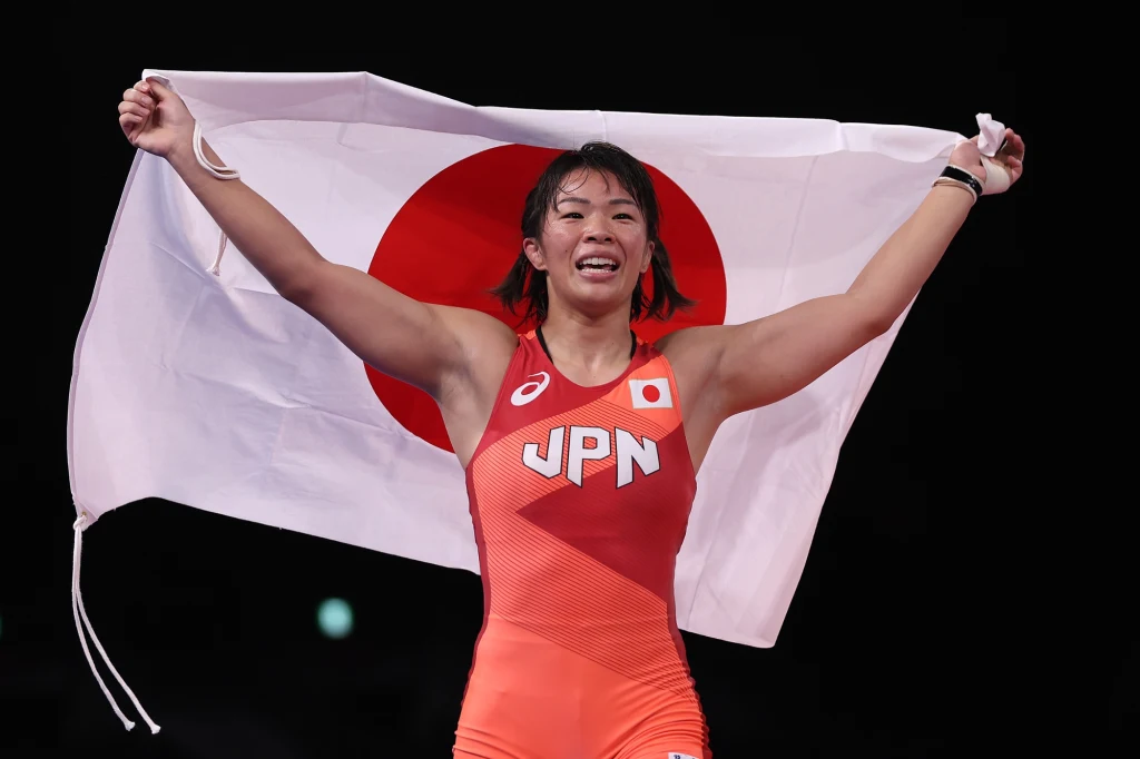 Zawodniczka w stroju sportowym z napisem JPN unosi flagę Japonii nad głową, wyraźnie świętując zwycięstwo i okazując radość