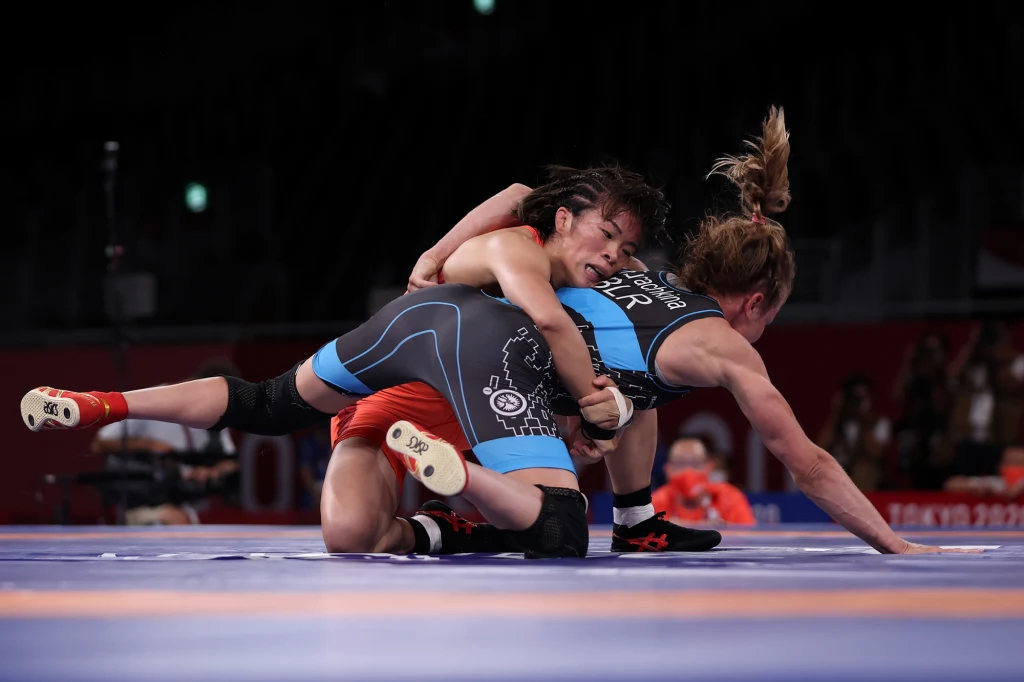 Dwie zawodniczki zmagają się na macie podczas meczu zapasów, jedna z nich stara się wykonać technikę obalenia, obie ubrane w profesjonalne stroje sportowe.