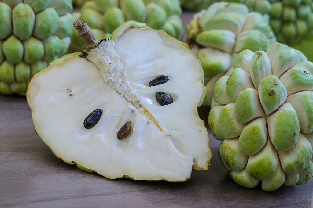 Rozkrojony owoc flaszowca na jasnym, drewnianym tle, z widocznym białym miąższem i czarnymi pestkami, w tle kilka całych owoców o zielonkawej skórce.