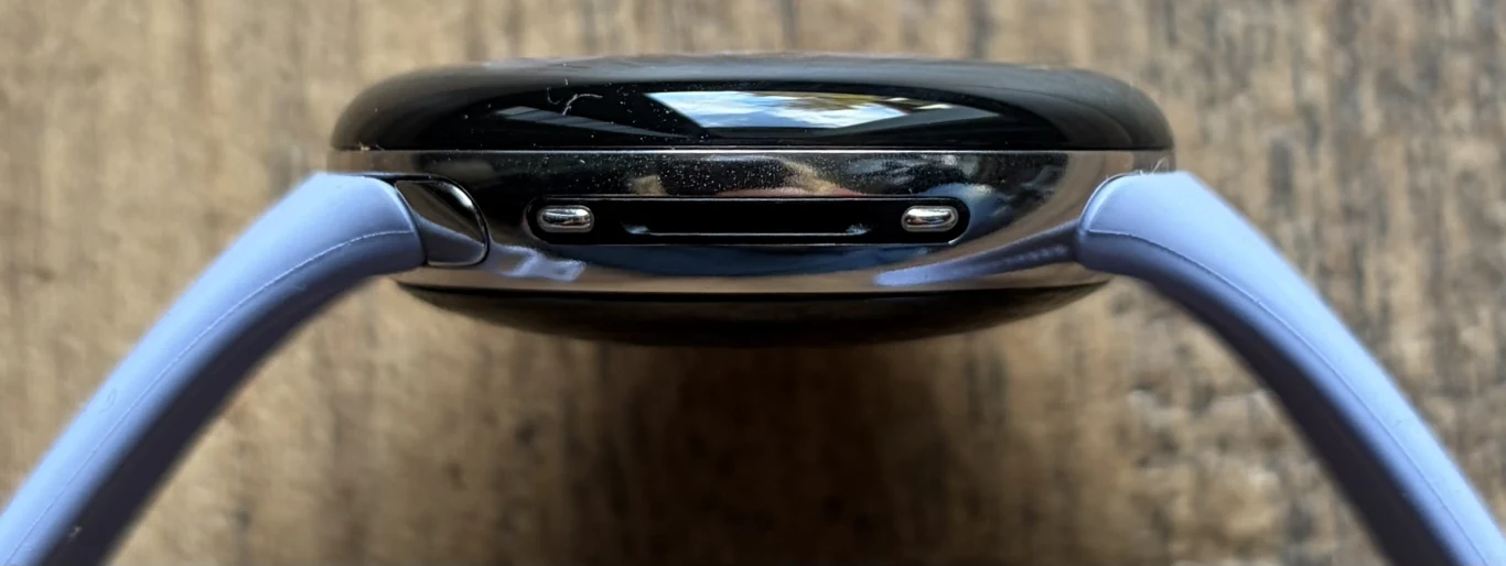 Google Pixel Watch 4 wprowadza pewne zmiany. Obejmują one głośniczek i piny dla ładowarki. Zbliżenie na spód czarnej koperty smartwatcha z fioletowym paskiem, widoczny port i przyciski boczne, tło drewniane.