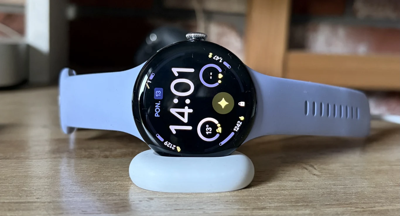 Googel Pixel Watch 4 dostarczany jest z nową ładowarką. Ładowanie jest szybsze i odbywa się w innej orientacji. Nowoczesny smartwatch z okrągłą kopertą na silikonowym pasku w jasnofioletowym odcieniu, wyświetlacz pokazuje godzinę 14:01 oraz różne wskaźniki fitnessowe i powiadomienia, urządzenie ustawione na białej, zaokrąglonej podstawce na drewnianym blacie.