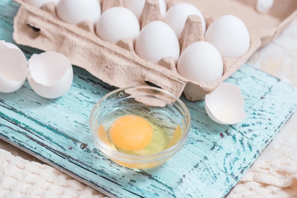 Segregujesz śmieci? Tego jednego błędu z opakowaniami po jajkach nie popełniaj Kilka białych jajek w kartonowym opakowaniu, jedno jajko rozbite i zawartość w szklanej miseczce, obok widoczne skorupki, wszystko na tle niebieskiej, przecieranej deski.