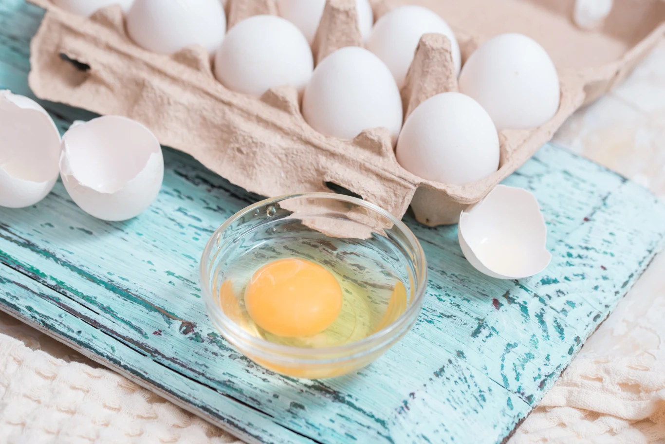 Kilka białych jajek w kartonowym opakowaniu, jedno jajko rozbite i zawartość w szklanej miseczce, obok widoczne skorupki, wszystko na tle niebieskiej, przecieranej deski.