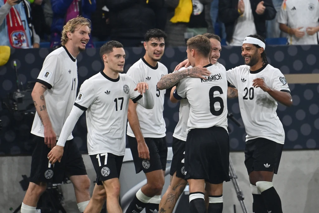 Reprezentacja Niemiec Grupa piłkarzy w białych koszulkach świętuje zdobycie bramki, przytulając się i uśmiechając, na tle trybun z kibicami.