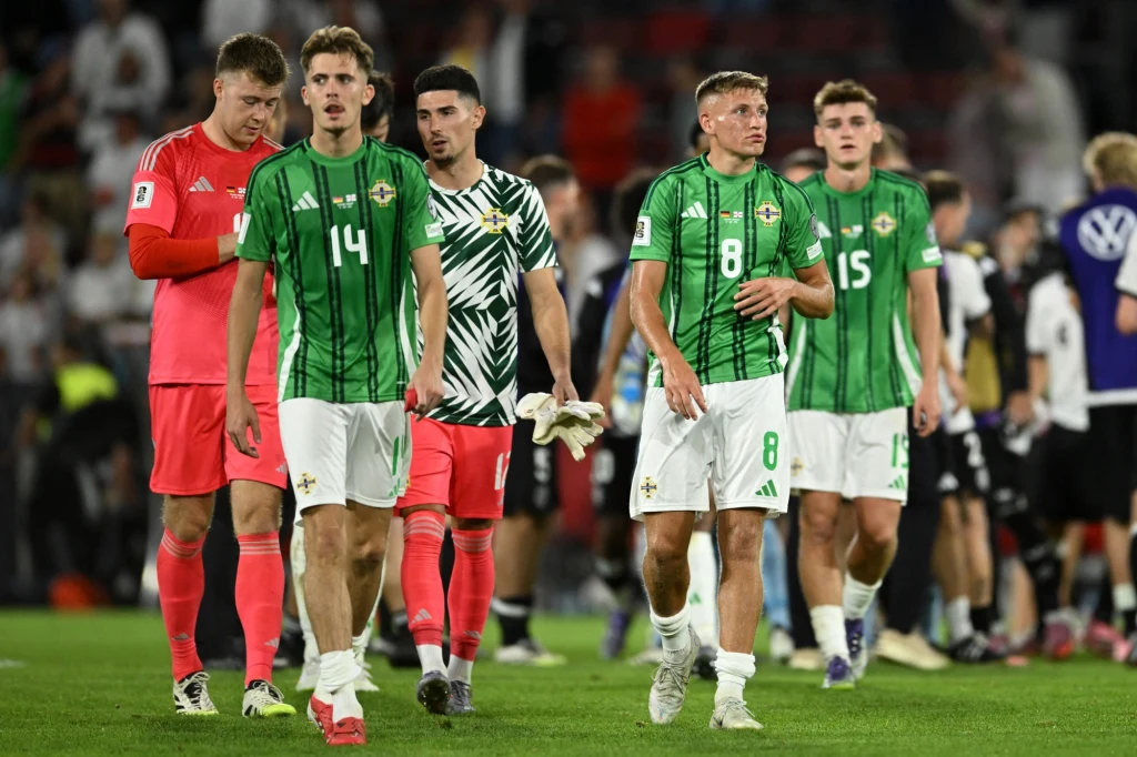 Piłkarze Irlandii Północnej Grupa piłkarzy w zielonych i białych koszulkach schodzi z boiska po zakończonym meczu, obok nich bramkarz w czerwonym stroju oraz zawodnicy w tle w czarnych i białych strojach, wokół stadionowy gwar.