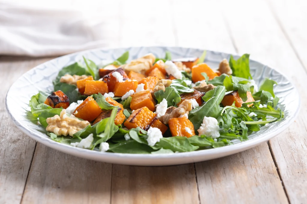 Zdrowa sałatka z dynią Sałatka z rukolą, pieczoną dynią, orzechami włoskimi i kawałkami sera feta podana na białym talerzu, ustawionym na jasnym drewnianym stole.
