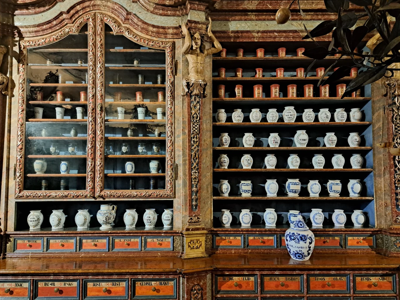 Bogato zdobiona drewniana szafa apteczna z licznymi ceramicznymi pojemnikami i szufladami oraz pojedynczym dużym dzbankiem w niebieskie wzory na blacie. Na półkach równo ustawione są białe i czerwone naczynia z napisami.