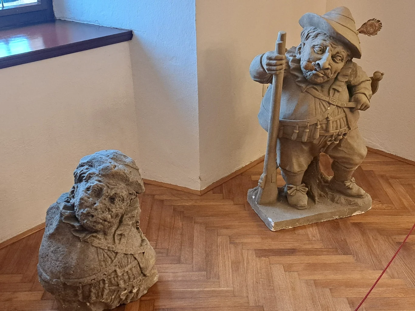 Dwie rzeźby kamienne ustawione w rogu pomieszczenia, jedna przedstawia postać siedzącą z wyraźnymi, realistycznymi rysami twarzy, druga to figura krasnoluda w kapeluszu z fajką, trzymającego laskę.