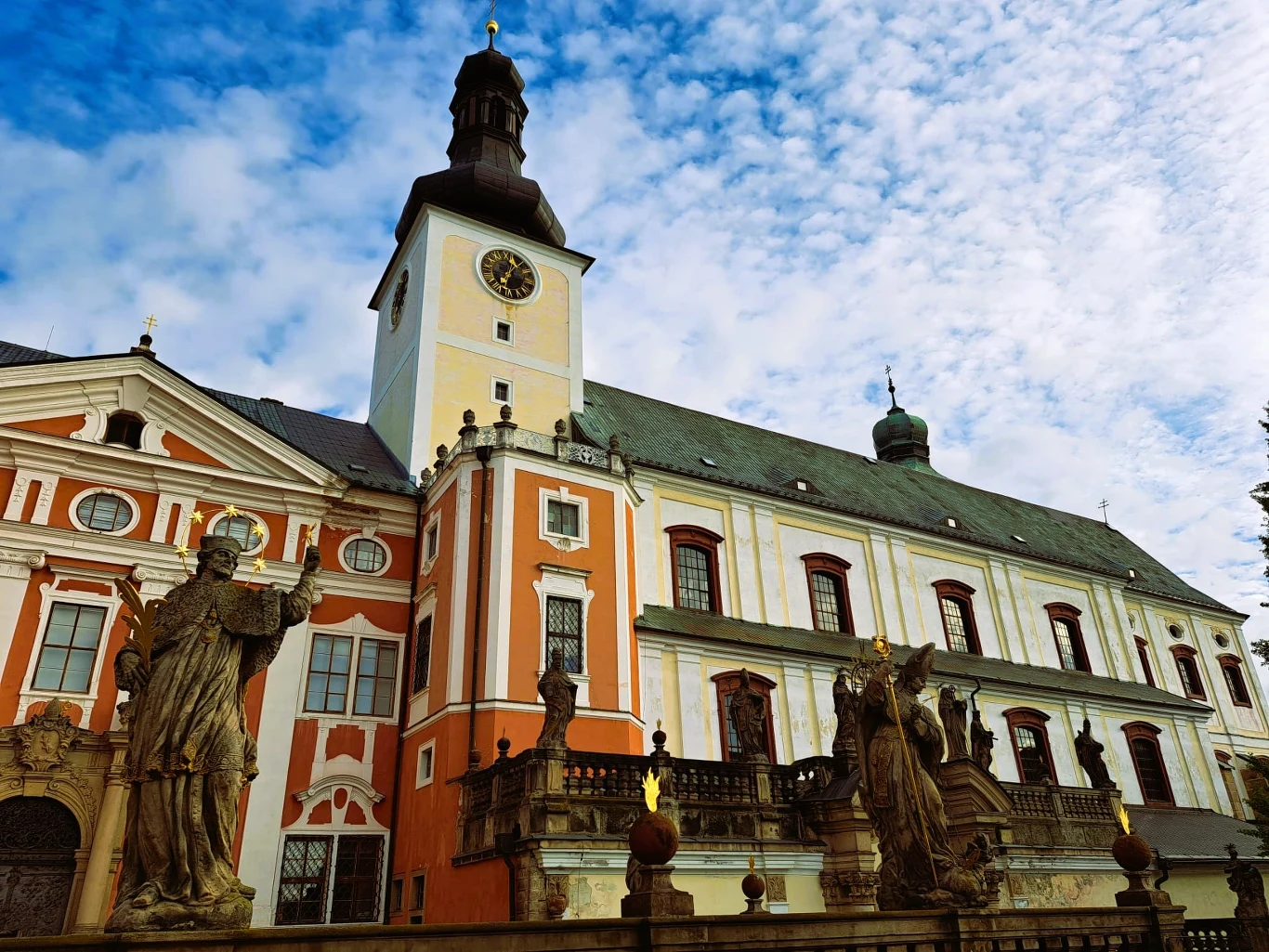 Barokowy kościół z jasnymi ścianami i wysoką wieżą zegarową, otoczony ozdobnymi rzeźbami świętych, na tle niebieskiego nieba pokrytego delikatnymi chmurami