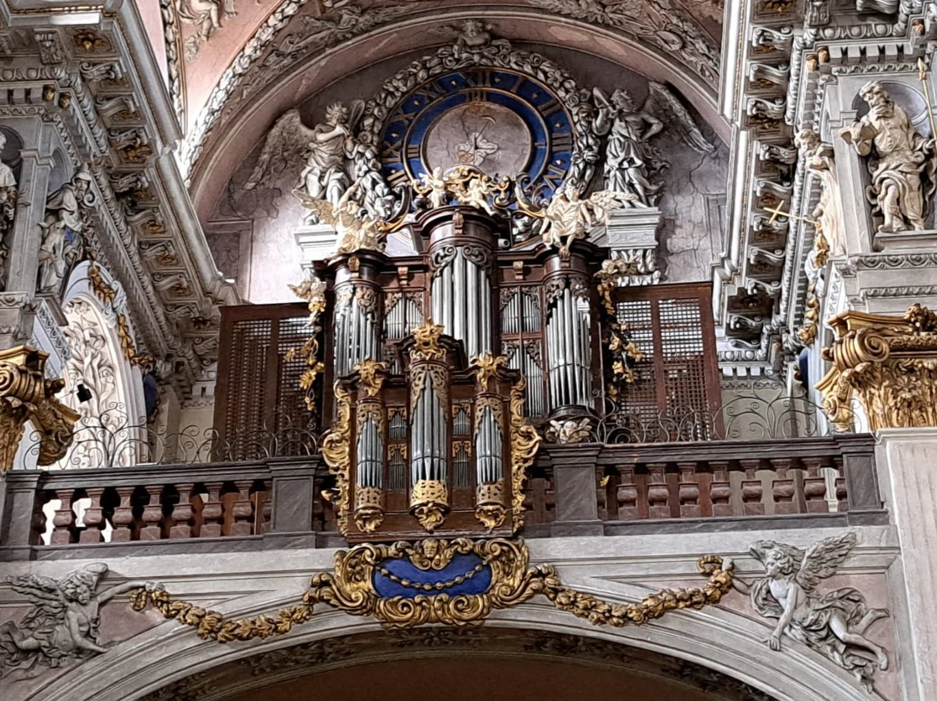 Barokowe organy umieszczone na dekoracyjnym chórze kościelnym, otoczone bogatymi złoceniami, rzeźbami aniołów oraz okazałym zegarem z niebiesko-złotą tarczą. Bogactwo detali i sztukaterii podkreśla monumentalny charakter wnętrza.