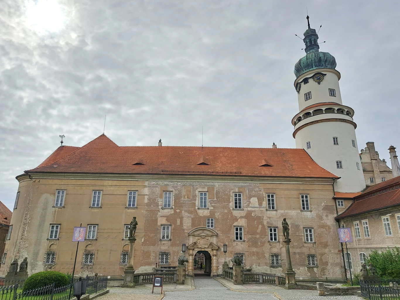 Frontowa fasada okazałego zamku z czerwonym dachem, licznymi oknami i zdobionym wejściem, obok wieża z zieloną kopułą i zegarem. Przed budynkiem znajduje się brukowany dziedziniec oraz figury kamienne.
