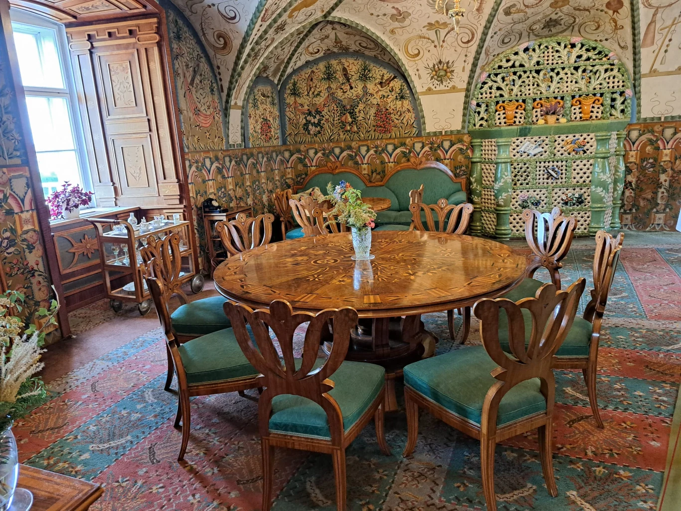Okrągły, drewniany stół z misternymi zdobieniami otoczony eleganckimi krzesłami z miękkim, zielonym obiciem, w centrum pomieszczenia o bogato dekorowanych ścianach i sklepieniu, z ozdobnym, zielono-białym kaflowym piecem w rogu oraz kolorowym, wzorzyst...