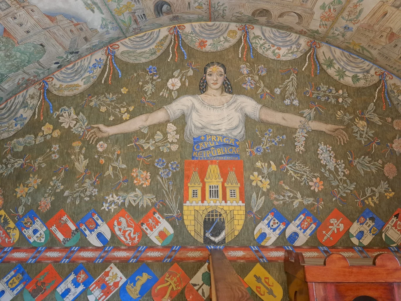 Kobieta z rozpostartymi ramionami otoczona kwiatami, nad którą umieszczono napis 'Praga Republika Czeska', poniżej widoczny herb z budynkiem, a całość dekorowana licznymi kolorowymi godłami i ornamentami.