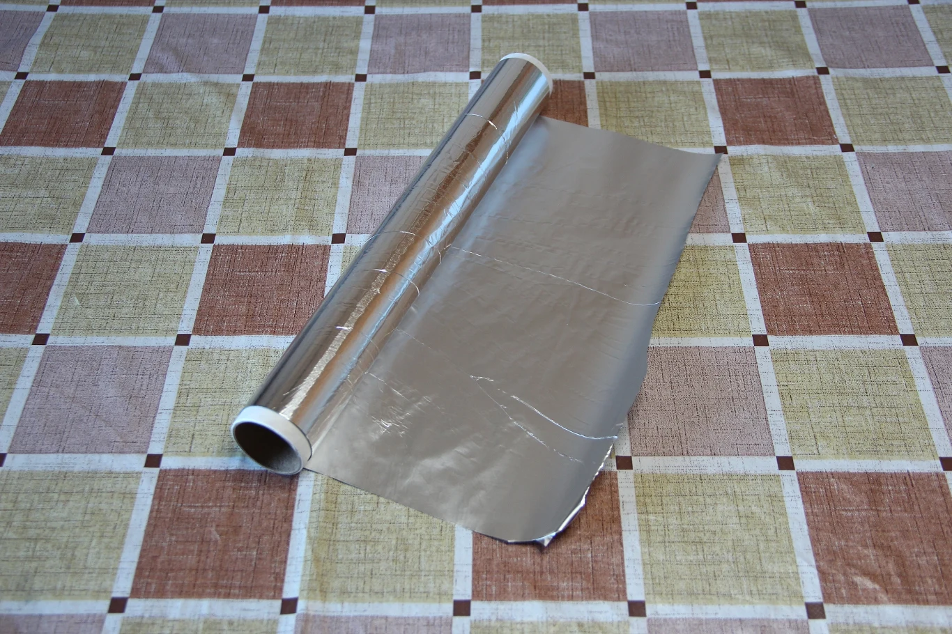 Rolka folii aluminiowej rozwiniętej na stole pokrytym obrusie w kratkę, z lekko postrzępionym końcem arkusza.