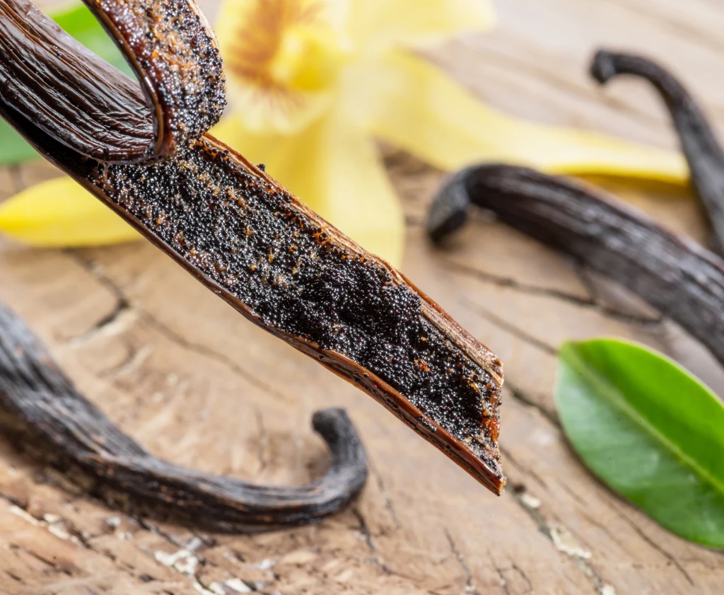 Laska wanilii przecięta wzdłuż, z widocznymi aromatycznymi ziarnami, na tle kwiatów wanilii i zielonych liści, umieszczona na drewnianym podłożu