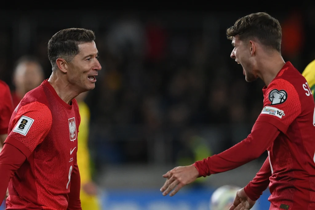 Dwóch piłkarzy w czerwonych koszulkach z orłem na piersi, ujętych w momencie radości na boisku podczas meczu piłkarskiego.