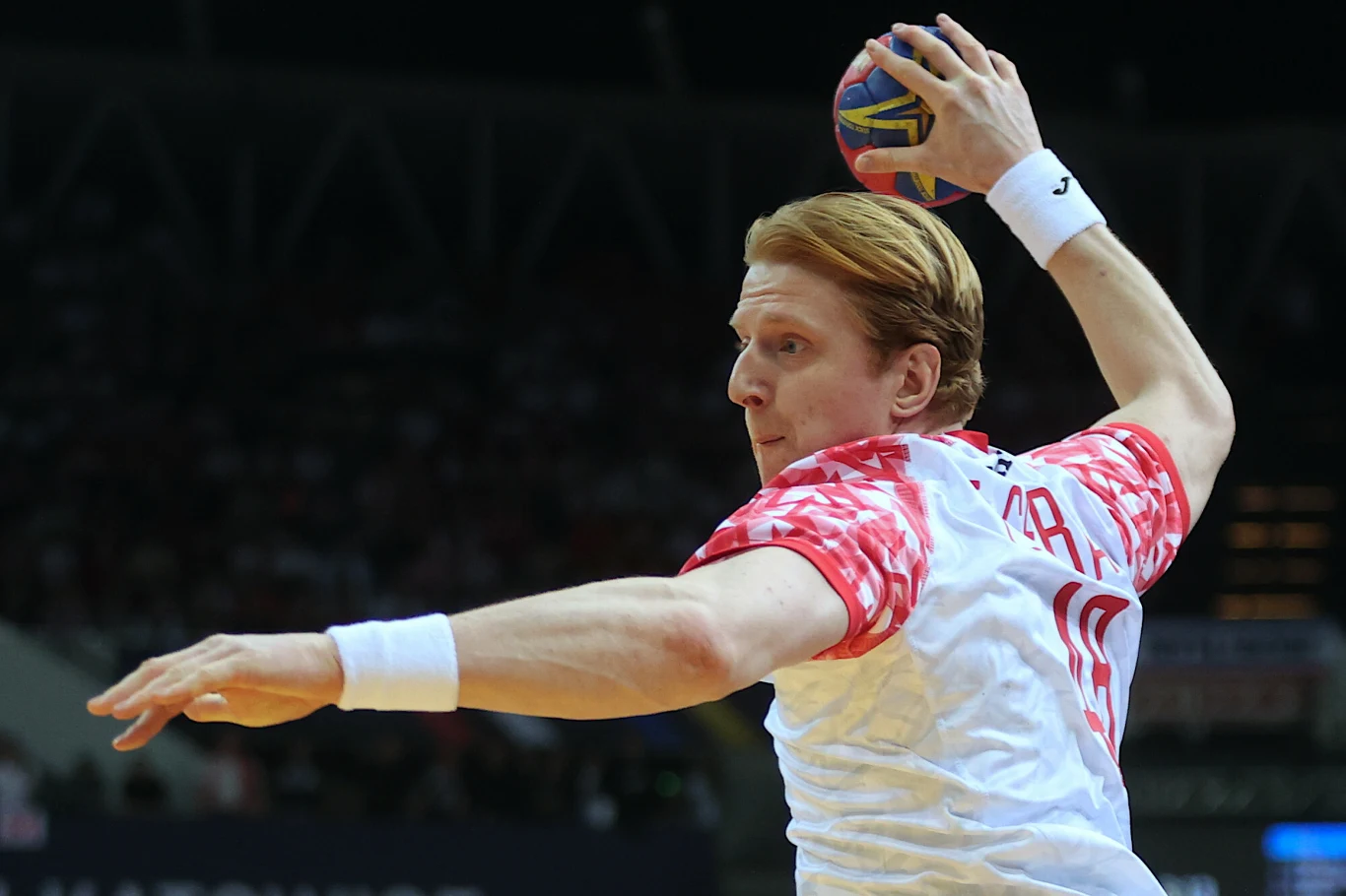 Tomasz Gębala Zawodnik w białej koszulce z czerwonymi rękawami trzyma piłkę do piłki ręcznej i przygotowuje się do rzutu, intensywnie skupiony na boisku, w tle niewyraźna widownia w hali sportowej.