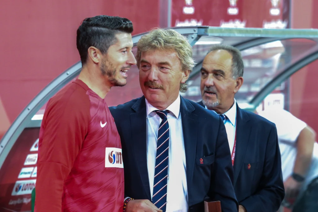 Trzech mężczyzn w eleganckich strojach sportowych i garniturach rozmawia ze sobą przy ławce rezerwowych na stadionie piłkarskim, atmosfera wydaje się przyjazna i profesjonalna.