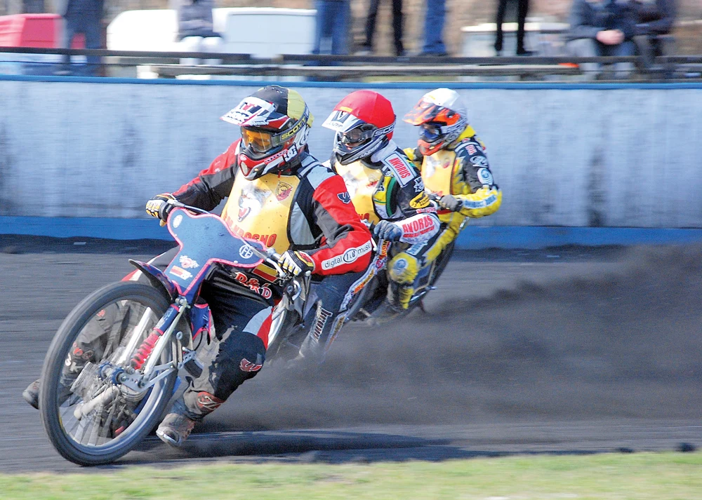 Krośnieński żużel przeszedł burzliwą drogę pełną wzlotów i upadków Trzech motocyklistów w kolorowych kombinezonach i kaskach rywalizujących podczas wyścigu żużlowego na torze, zbliżają się do zakrętu w trakcie jazdy po czarnej nawierzchni, z motocykli unosi się kurz.