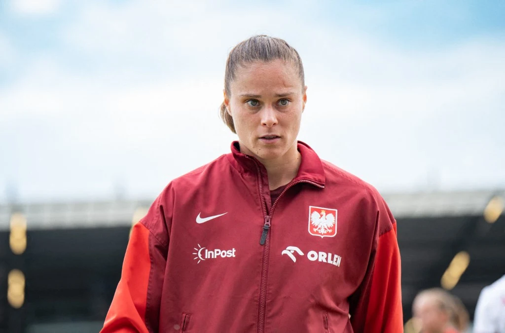 Osoba w czerwonej sportowej kurtce z widocznym orłem na piersi i logotypami sponsorów stoi na tle rozmytego stadionu, patrząc poważnie przed siebie.