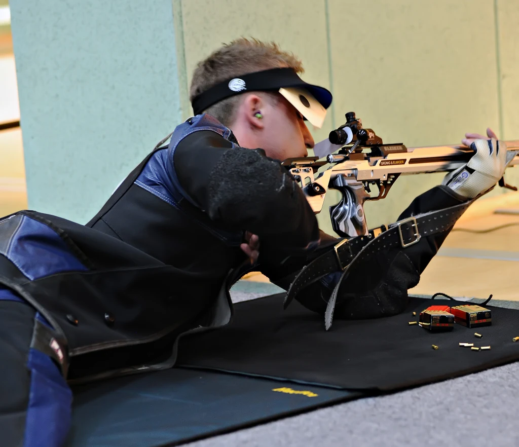 Sportowiec w stroju ochronnym leży na macie i celuje z karabinu sportowego, obok leży amunicja. Sytuacja odbywa się wewnątrz strzelnicy.