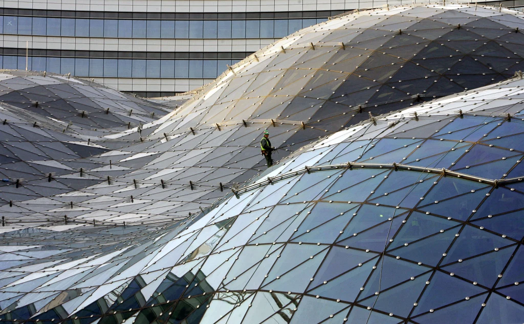 Pracownik w kombinezonie zabezpieczającym porusza się po dużym szklanym dachu o futurystycznej, falistej konstrukcji złożonej z licznych tafli szkła i metalowych łączeń, w tle widoczne jest nowoczesne biurowe oszklenie.