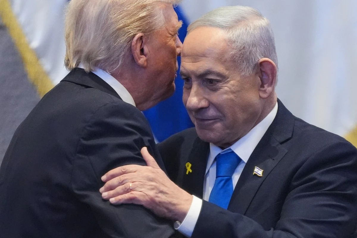 Prezydent Stanów Zjednoczonych Donald Trump i premier Izraela Binjamin Netanjahu Dwóch mężczyzn odzianych w garnitury w uścisku dłoni, jeden z nich szepcze coś do ucha drugiego, który patrzy poważnie do przodu, na klapie ma żółtą wstążkę.
