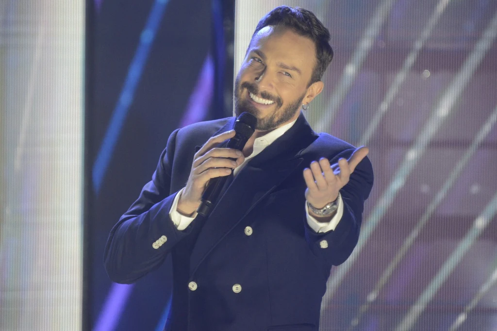 Sławomir Uniatowski Elegancko ubrany mężczyzna z mikrofonem w dłoni, uśmiecha się i gestykuluje dłonią podczas występu na scenie z jasnym, rozmytym tłem efektów świetlnych.