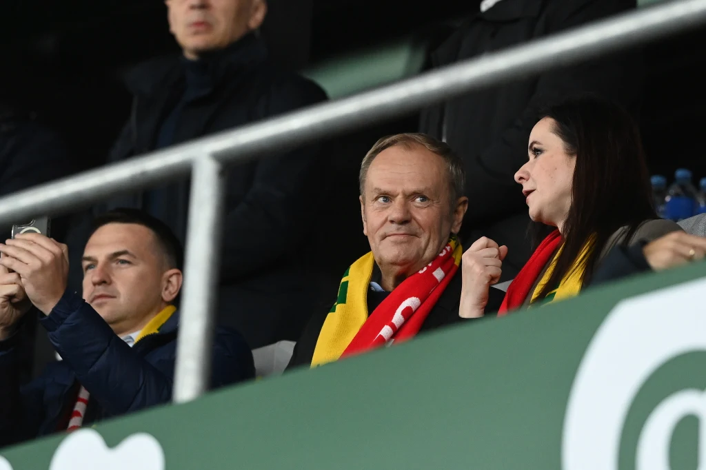 Premier Donald Tusk i szefowa litewskiego rządu Inga Ruginiene na meczu w Kownie Trzy osoby siedzą na trybunach stadionu, dwie z nich ubrane w szaliki kibicowskie, jedna z kobiet rozmawia gestykulując ręką, mężczyzna obok patrzy na nią uważnie, trzecia osoba fotografuje coś telefonem.