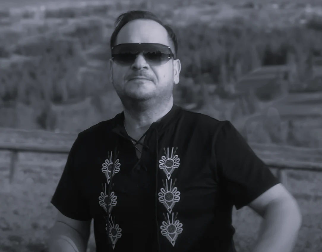 Ben Vito w teledysku "Góraleczka mała" Mężczyzna w ciemnych okularach przeciwsłonecznych i czarnej koszuli z wyszywanym wzorem stojący na tle rozmytego krajobrazu z drzewami i ogrodzeniem.