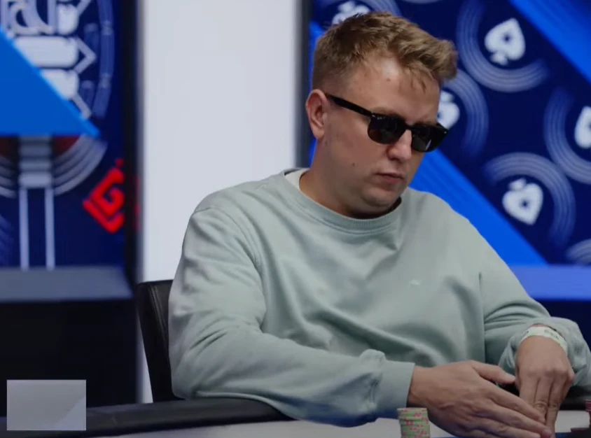 Tomasz Brzeziński Mężczyzna w jasnej bluzie z długim rękawem i ciemnych okularach przeciwsłonecznych siedzi przy stole pokerowym, skupiając się na żetonach i kartach, w tle widoczne elementy dekoracyjne związane z grą w pokera.