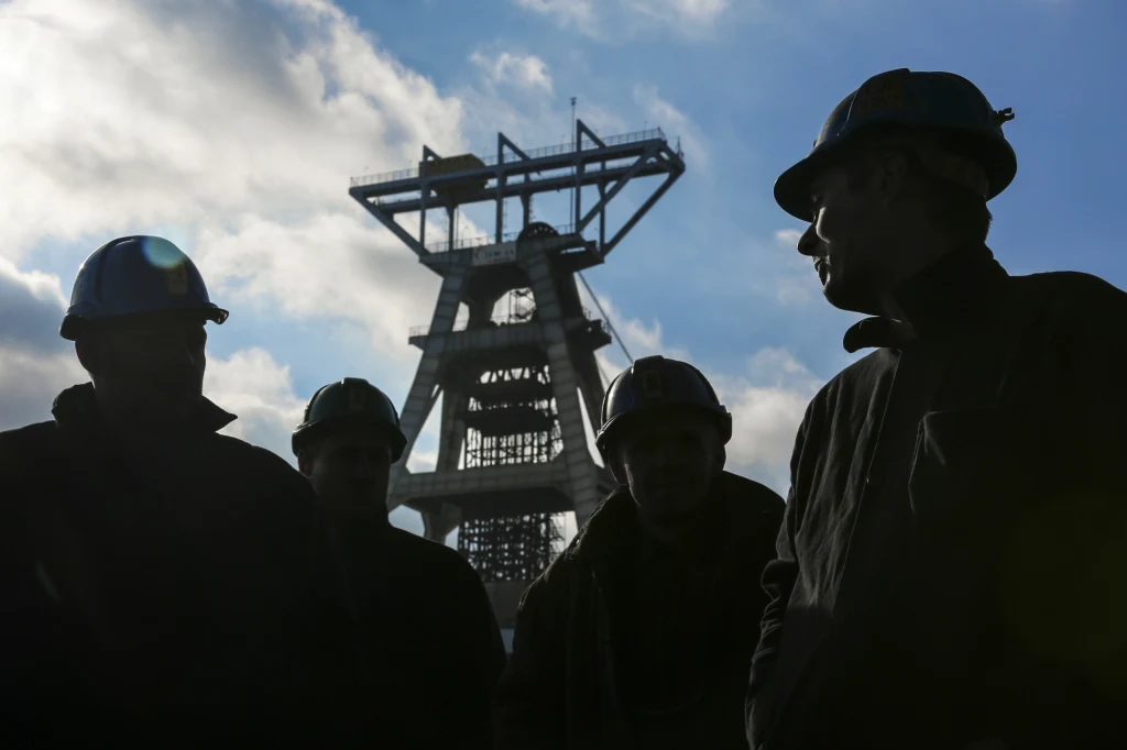 Sylwetki kilku górników w kaskach ochronnych na tle szybu kopalnianego oraz nieba pokrytego chmurami, w tle wyraźnie widoczna stalowa konstrukcja szybu.