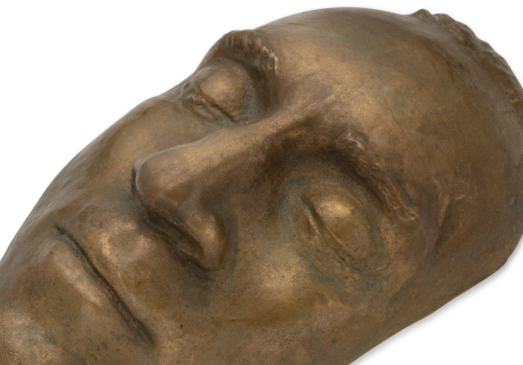 Brązowy odlew pośmiertnej marki Włodzimierza Wysockiego w zbiorach domu aukcyjnego Hermitage Fine Art Metalowa maska pośmiertna przedstawiająca twarz mężczyzny z zamkniętymi oczami, wykonana z brązu, eksponująca szczegóły rysów twarzy i strukturę materiału.