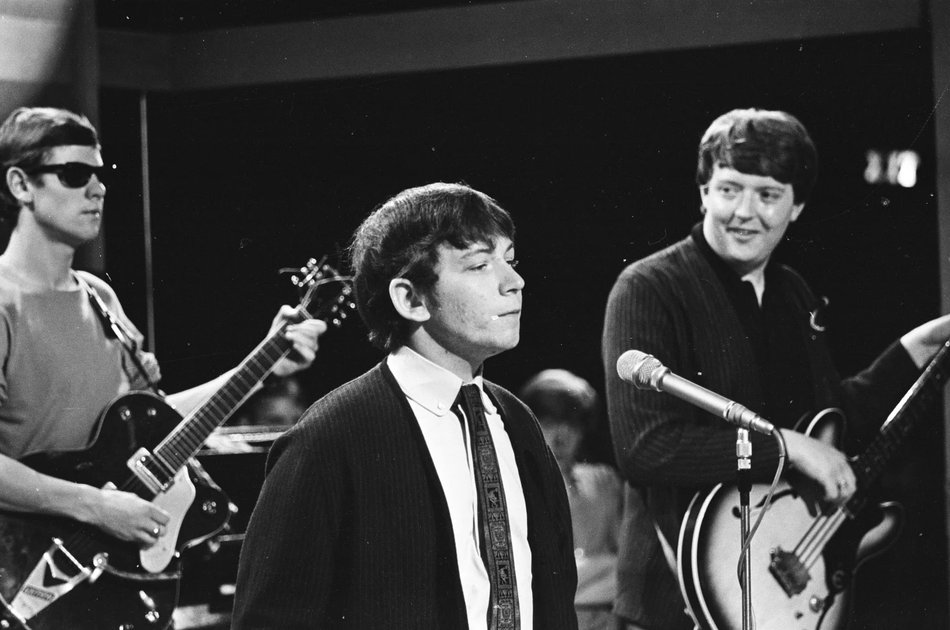 Hilton Valentine, Eric Burdon i Chas Chandler z The Animals Trzech mężczyzn na scenie muzycznej, środkowy z nich stoi przy mikrofonie, pozostali grają odpowiednio na gitarze i gitarze basowej, w tle publiczność.