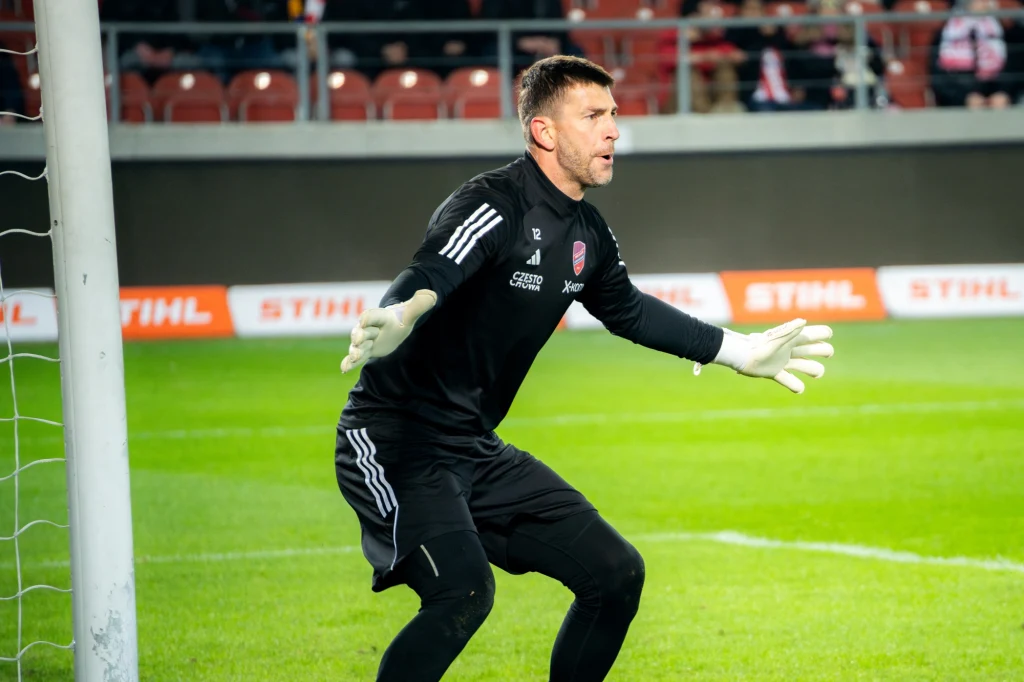 Bramkarz piłkarski w czarnym stroju przygotowuje się do obrony na tle bramki podczas meczu na stadionie, wokół widoczne trybuny z kibicami.